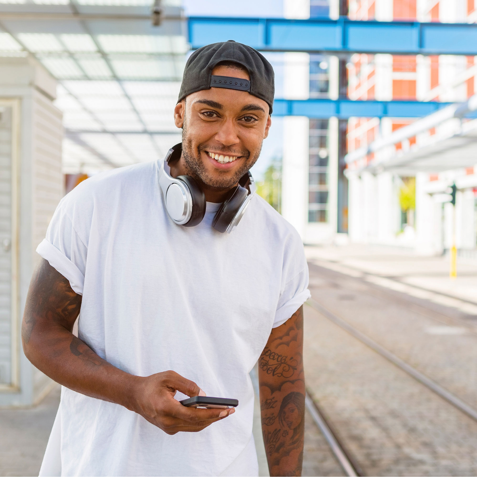 leende unga man med hörlurar och smartphon väntar vid spårvagnshållplatsen